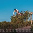 Ein Fahrradfahrer springt in der Luft vor einem blauen Himmel. Unten ist ein Kalender mit Zahlen und Wochentagen.