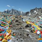 Bunte Gebetsfahnen flattern auf einem felsigen Gebirgspass. Im Hintergrund schneebedeckte Berge unter klarem Himmel.