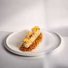 Keine sichtbaren Texte. Ein kunstvoll angerichtetes Gericht mit einer Wurst, dekoriert mit Blüten und Chips, auf weißem Teller.