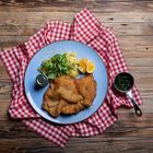 Ein Teller mit Schnitzel, Kartoffelsalat, Feldsalat, Zitronenscheiben, auf einem rot-weiß karierten Tuch.