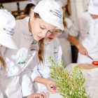 "Europa Miniköche" auf weißen Kappen. Kinder in Kochkleidung bereiten Essen zu, mit Kräutern auf dem Tisch.