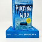 Ein blaues Buch mit „Pucking Wild“ von Emily Rath, „Der spicy TikTok-Erfolg“ im gelben Kreis, auf Holzpier-Hintergrund.