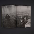 Keine erkennbaren Texte. Drei Taucher in dunklen Anzügen stehen im Wasser, einer hebt einen großen, nassen Gegenstand.