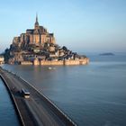 Eine große Abtei auf einer felsigen Insel, verbunden durch einen Damm, umgeben von Wasser und leichter Bewölkung.