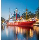 "Deutsche Bucht" auf einem roten Schiff, angedockt am Wasser vor einem städtischen Hintergrund, abends beleuchtet.