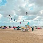 Himmel mit Wolken, Menschen am Strand beim Kitesurfen, bunte Kites am Boden und in der Luft, Meer im Hintergrund.