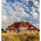 Kurhaus in den Dünen von Zingst. Fachwerkhaus mit rotem Dach, umgeben von hohem Gras. Blauer Himmel mit vielen Wolken.