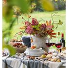 Text: "42 OKTOBER", "MO 18 DI 19 MI 20 DO 21 FR 22 SA 23 SO 24". Garten mit gedecktem Tisch, Blumen, Wein, Käse und Obst.