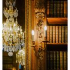 Text: "43 OKTOBER MO 25 DI 26 MI 27 DO 28 FR 29 SA 30 SO 31" 
Ein opulenter Kronleuchter spiegelt sich in einem vergoldeten Spiegel; daneben eine antike Bücherwand.