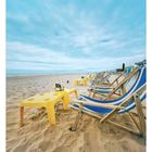 Liegestühle und gelbe Tische am sandigen Strand unter bewölktem Himmel, mit Meerblick im Hintergrund.