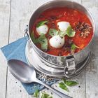 Weißer Topf mit roter Tomatensuppe, Mozzarella-Kugeln und Basilikumblättern auf einem blauen Serviettendeckel.