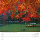 "Fluss Katsura in Arashiyama in Kyōto, 41. Woche, Oktober." Leuchtend rote und orange Blätter über grünem Fluss. Boot im Wasser.