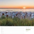 42. Woche Oktober 2027. Weiße Strandkörbe am Sandstrand mit Sonnenuntergang über dem Meer, Gras im Vordergrund.