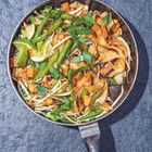 Breite Nudeln, Gemüse, grüner Lauch, Limettenscheiben und Tofu in einer metallischen Pfanne auf grauer Oberfläche.