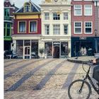 Texte im Bild: "Bolla.com", "ZANZZ". Drei historische, bunte Häuser, Steinpflaster. Fahrradfahrer rechts.