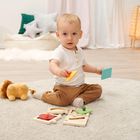 Ein Kleinkind sitzt auf einem Teppich und spielt mit buntem Holzspielzeug, im Hintergrund sind Kissen und ein Plüschtier.