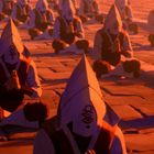 Männer mit Kapuzen und Augen-Symbolen meditieren im Sonnenuntergang auf einem gepflasterten Platz.