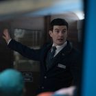 Ein Mann in Uniform gestikuliert in einem Innenraum, umgeben von diffusen, dunklen Farben. Schilder im Hintergrund.