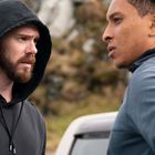 Zwei Männer stehen sich draußen gegenüber, einer trägt einen schwarzen Kapuzenpullover, der andere einen dunkelblauen.