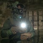 Eine Person in Schutzanzug und Gasmaske steht in dunklem Wasser mit einer leuchtenden Taschenlampe.