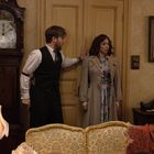 Ein Mann und eine Frau stehen vor einer Holztür in einem Wohnzimmer mit altmodischer Tapete, einer Stehlampe und einem großen Wandteppich.