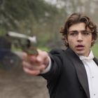 Ein junger Mann in einem eleganten Anzug hält eine Pistole und schaut entschlossen in die Ferne.