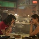 Im Hintergrund steht "EXIT". Zwei Personen unterhalten sich in einem gemütlichen Raum. Eine trägt ein rotes T-Shirt, die andere ein ärmelloses Top.