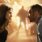 Ein Mann und eine Frau schauen sich intensiv an. Im Hintergrund ist eine verschwommene, helle Explosion erkennbar.