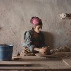 Eine Frau töpfert an einer Drehscheibe. Sie trägt ein kariertes Hemd und ein pinkes Tuch im Haar, umgeben von Werkzeugen.