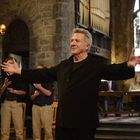 Ein Mann breitet die Arme aus, umrahmt von singenden Jungen in einer Kirche mit Buntglasfenstern und Kerzenleuchter.