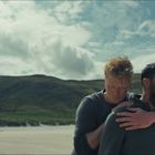 Zwei Männer am Strand, einer hält den anderen, der laut schreit. Im Hintergrund sind Hügel und ein Haus.