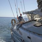 Ein Mann steuert ein Segelboot auf offenem Meer. Das Segel ist teilweise eingerollt. Es ist ein klarer, sonniger Tag.