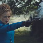 Eine Frau in einem blauen Mantel schießt mit einer Pistole. Rauch steigt auf, und die Umgebung ist grün und bewaldet.