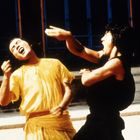 Zwei Männer kämpfen: Einer in Gelb mit erhobenem Arm, der andere in Schwarz schlägt zu. Eine Treppe im Hintergrund.