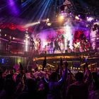 Leuchtstäbe, Partyatmosphäre, Bühne mit Tänzern, farbige Lichter, Publikum jubelt, Festlicher Raum mit aufregender Stimmung.