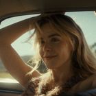 Ich kann den Text nicht erkennen. Eine Frau sitzt lächelnd im Auto und hält die Arme hoch, Sonnenlicht fällt auf sie.