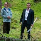 Ich kann keine Texte sehen. Zwei Männer stehen im Wald; einer trägt einen Anzug und hält eine Schaufel.