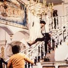 Ich kann den Text nicht erkennen. Eine Person schwingt an einem Kronleuchter über eine prächtige Treppe. Im Hintergrund ein Wandgemälde.