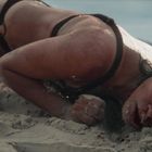 Es sind keine Texte zu erkennen. Eine Frau liegt erschöpft am Strand, der Kopf im Sand, Meer im Hintergrund.