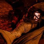 Eine Person in einem Kapuzenmantel mit einer bedrohlich bemalten Maske im rötlichen Licht, in einer dynamischen Bewegung.
