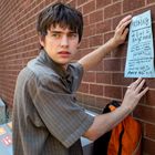 "MISSING" und "Digits of Emily’s number" stehen auf einem Zettel. Ein junger Mann klebt den Zettel an eine Backsteinmauer.