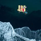 Zwei Personen liegen auf bunten Handtüchern im Wasser. Im Vordergrund ist Schaum von anrollenden Wellen zu sehen.