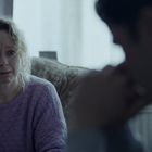 Eine Frau in einem lila Pullover wirkt traurig oder besorgt, während sie einem Mann gegenübersitzt. Im Hintergrund ist ein Vorhang.