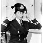 Eine Frau in Uniform mit doppelreihiger Jacke steht vor einem Spiegel und richtet ihren Hut. Am Hut ist ein maritimes Abzeichen.