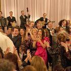 Eine Feier mit vielen Menschen. Eine Braut und ein Bräutigam werden auf Stühlen hochgehoben, während die Menge jubelt und klatscht.