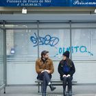 Aufschrift: "Plateaux de Fruits de Mer, Poissonnerie Jean". Mann und Frau sitzen in einer Bushaltestelle, sie unterhalten sich.
