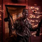 Ein Mann in verschmutzter Kleidung hält zwei Gewehre, die Holzwand im Hintergrund ist mit Einschusslöchern übersät.