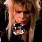 Ein Mann mit wildem blondem Haar schaut konzentriert in eine Kristallkugel, die sein Gesicht verzerrt widerspiegelt.