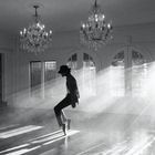 Keine Texte sichtbar. Eine Person mit Hut steht auf Zehenspitzen in einem sonnenbeschienenen Raum mit Kronleuchtern.