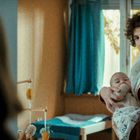 Keine sichtbaren Texte. Ein Mann in orangefarbener Strickjacke hält ein Baby in einem Raum mit Fenstern und blauen Vorhängen.